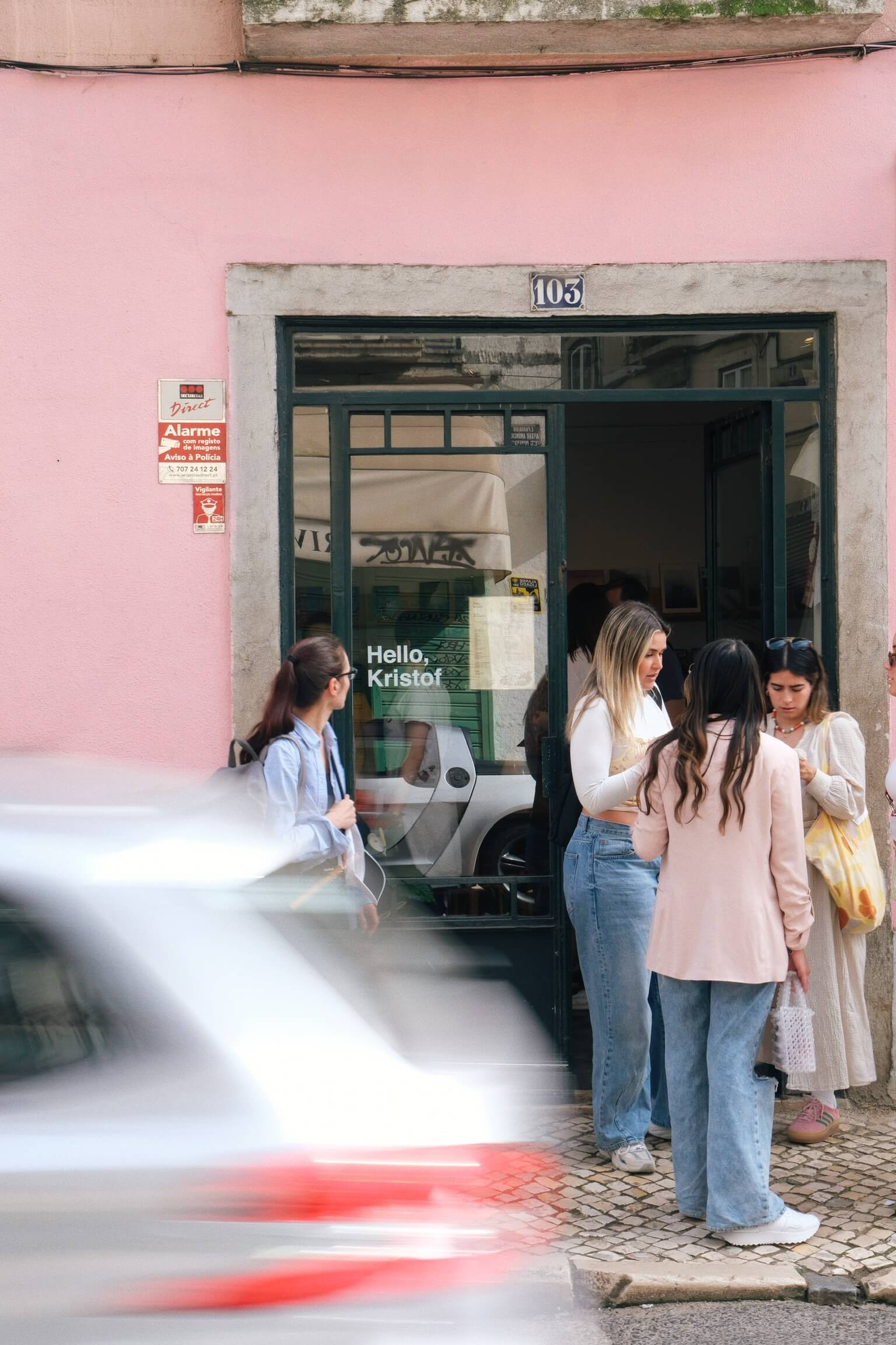 People standing outside the entrance of Hello, Kristof coffee shop in Bica, with a pink building facade and a car passing